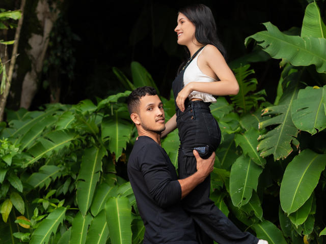 EimyAndMaxi with ass raised, wearing black pants and white top, held by EimyAndMaxi with dark hair, black shirt, and slight beard, in lush green jungle.