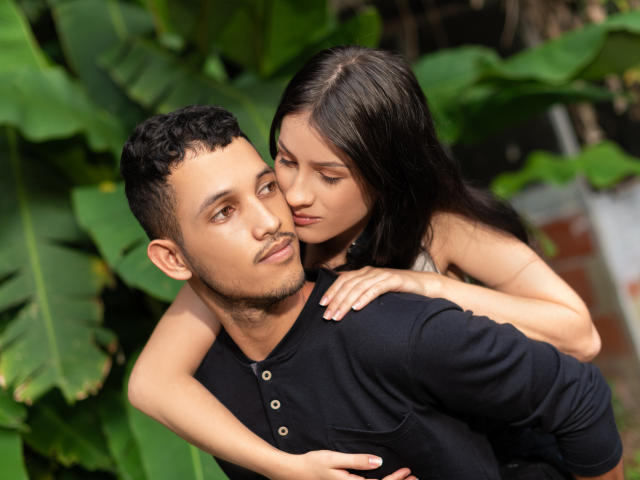 EimyAndMaxi with dark hair and pale skin, nose to nose with EimyAndMaxi, short curly brown hair, tan skin, black shirt, arms around each other, green leafy background.