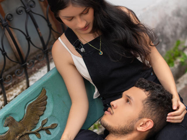EimyAndMaxi with dark hair, black sleeveless top, and white shirt, leans over Maxi with curly brown hair, black shirt, looking up. She's touching his head, he gazes up.