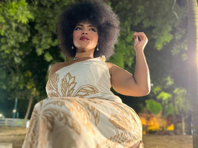 AliceJelanni with big Afro, wearing a white dress with gold patterns, holding her hair up, smiling, standing outdoors at night, light from the right, showing her ass.