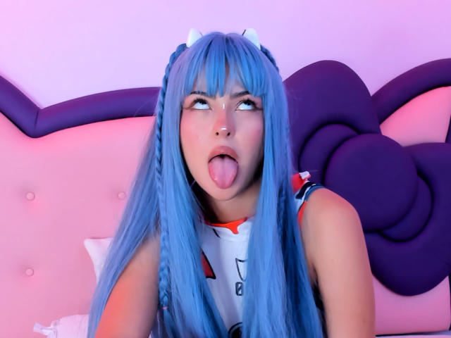 RheaVolk, con su largo cabello azul, sacando la lengua, vestida con un traje blanco y rojo con orejas de gato, tiene un piercing en la nariz y mira hacia arriba con una expresión juguetona..