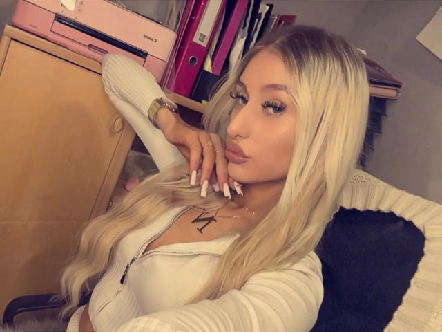 NatalieSusse-hot with long blonde hair, wearing a white zip-up top, resting her chin on her hand, showing off her large boobs and long, white-painted nails.