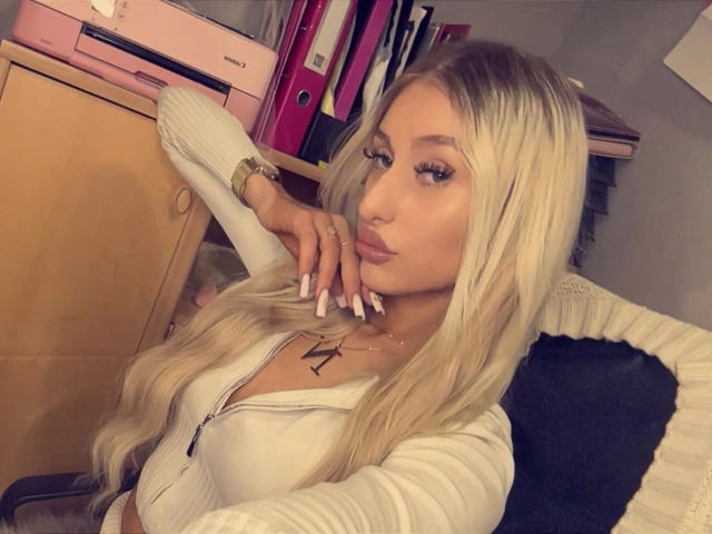 NatalieSusse-hot with long blonde hair, full lips, and large boobs, wearing a white zip-up top, leaning on a desk, gold wristwatch, and pink ring binder in the background.