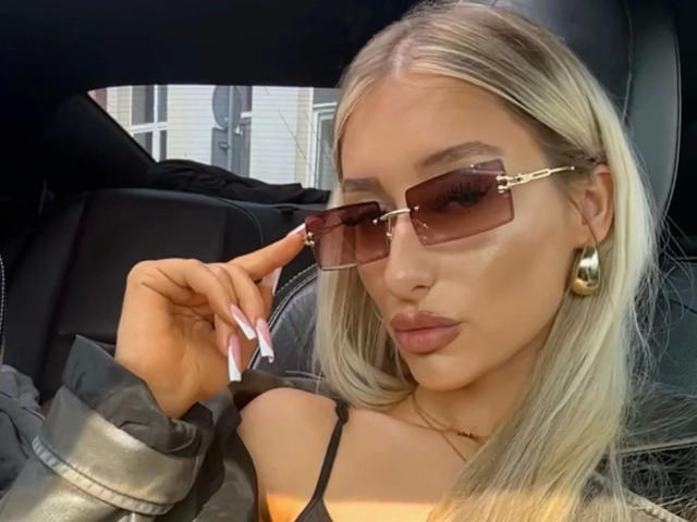NatalieSusse-hot, blonde, wearing gold earrings, brown sunglasses, and a grey jacket, with her pink-nailed hand touching her face, showing off her glossy, full-ass lips.