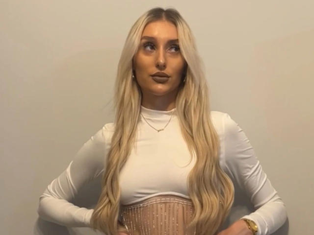 NatalieSusse-hot has long blonde hair, wears a white long-sleeve top, and a beige corset, looking to the side with hands on her hips, wearing a gold watch.