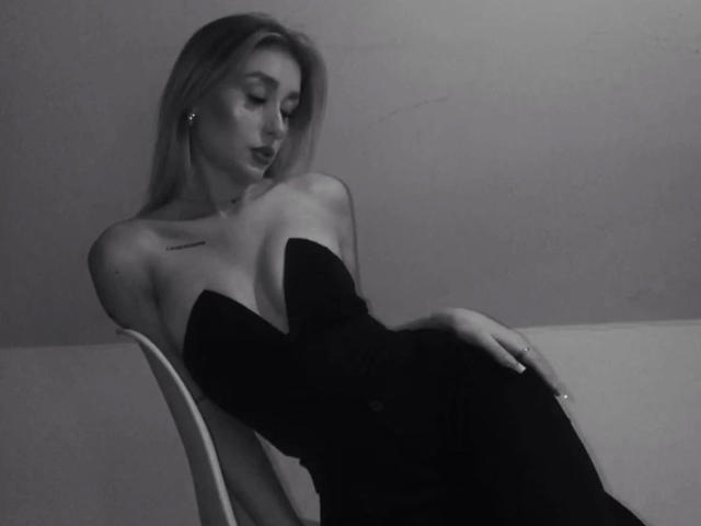 NatalieSusse-hot with her tits barely covered in a black strapless dress, sitting on a chair, looking down with a relaxed, sultry expression in a grayscale photo.