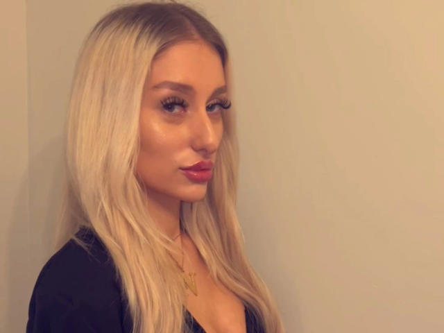 NatalieSusse-hot with blonde hair, long eyelashes, and red lipstick, wearing a black top, looking to the side with a slight pout, and a small gold necklace.