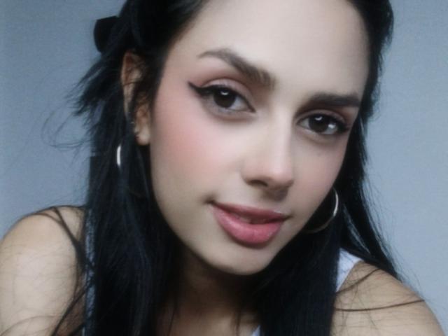 AnaisHilton has black hair, thick eyebrows, winged eyeliner, pink lipstick, and a slight smile, wearing a white tank top with large silver hoop earrings.
