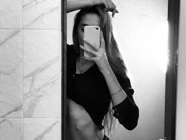 Black-and-white selfie of EviLune with long hair, holding phone, covering face, wearing a lifted shirt, showing a flat stomach and a small, visible tattoo on her side.