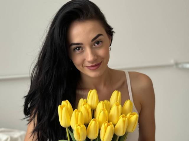 EviLune with long black hair, yellow tulips in front, smiling softly, wearing a white tank top, showing a cute face and small tits, against a plain white wall.