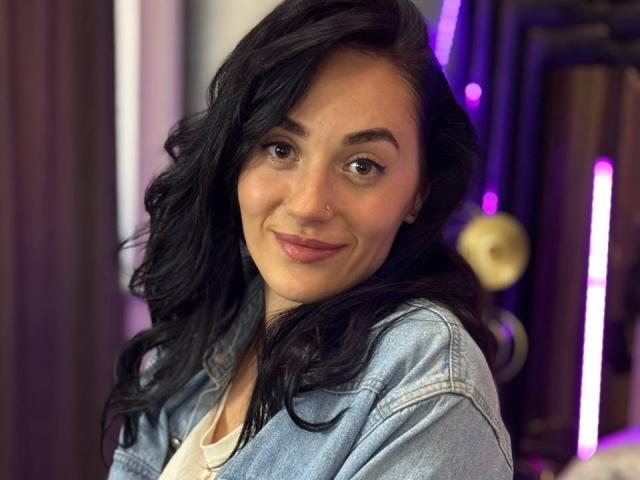 EviLune with black wavy hair, light brown eyes, and pink lipstick, wearing a blue denim jacket, smiling softly, showing subtle makeup, and a small nose ring.
