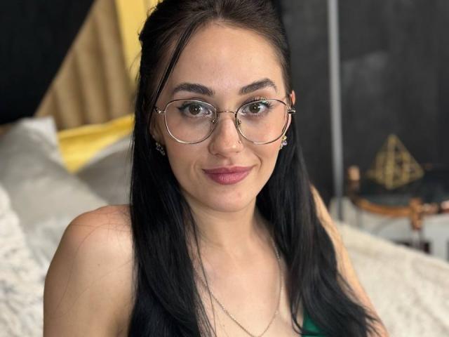 EviLune with long black hair, glasses, and pink lipstick, smiling at the camera, showing off her cute face and small tits. She's wearing a green top and a necklace.