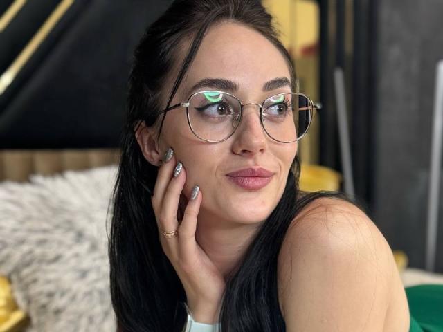EviLune with black hair, glasses, and pink lipstick, touching her cheek, showing off her nails with a ring, looking at the camera with a slight smile.