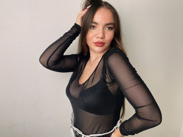 Kris Jenner, con cabello largo y castaño, vistiendo una blusa negra transparente de manga larga que deja ver sus senos, de pie frente a una pared lisa, con una mano en el cabello, mirando a la cámara..