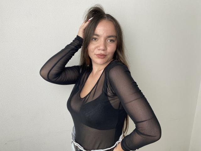 KrisTeigen with long brown hair, wearing a sheer black long-sleeve top, exposing small tits and a black bra, standing against a white wall, hand in hair.