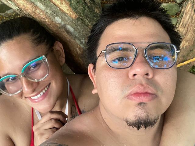 JuliaxRosee with blue-framed glasses, smiling, has boobs and a red strap, while a chubby guy with black hair and glasses, has a goatee, both in a selfie.