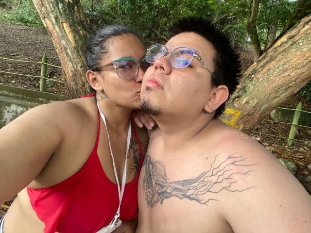 JuliaxRosee with dark hair, glasses, and red bikini top kisses a shirtless JuliaxRosee with tattoos, glasses, and short black hair. Both have medium skin and relaxed expressions.