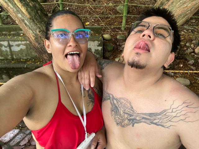 JuliaxRosee with big tits in a red bikini, tongue out, and blue glasses. JuliaxRosee's tattooed, shirtless boyfriend with short black hair and glasses, tongue out. Outdoor selfie.