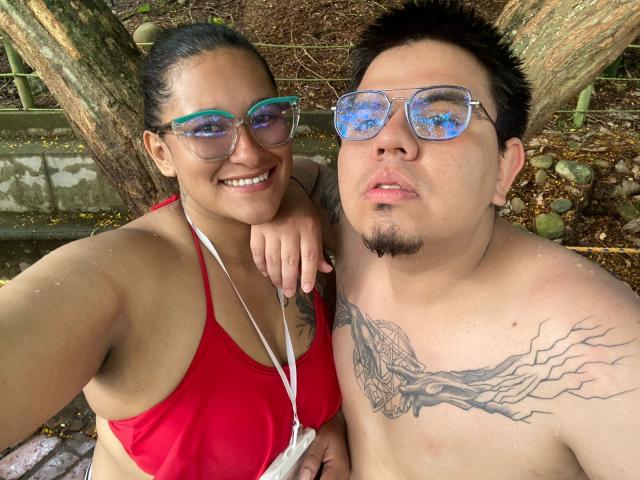 JuliaxRosee with green-tinted glasses, red bikini top, and tan skin, and a tattooed, shirtless man with blue-tinted glasses, and a small goatee, standing outdoors.