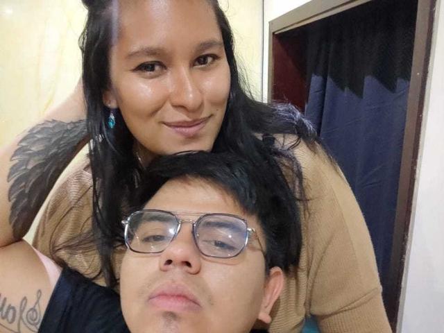 JuliaxRosee with black hair and tan top, leaning over a guy with glasses, black hair, and brown shirt, showing a small tattoo on her arm. Her tits are slightly visible.