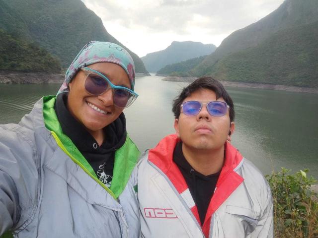 JuliaxRosee and a dude with dark hair, wearing blue sunglasses and a red and white jacket, standing by a river, smiling. JuliaxRosee has a teal headscarf and green hoodie.