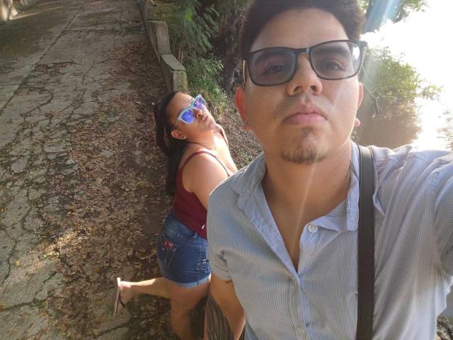 JuliaxRosee with blue reflective sunglasses, red top, denim shorts, and flip-flops, standing behind a guy in a light blue shirt and black suspenders, on a cracked stone path.