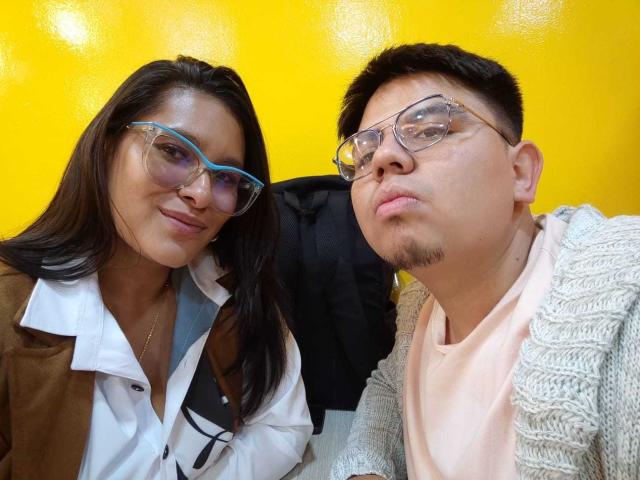 JuliaxRosee with glasses and brown jacket, next to a guy with glasses and a beige sweater, yellow background, both smiling. Boobs not visible. Ass not visible. Tits not visible.