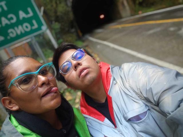 JuliaxRosee with teal glasses, black hair, green jacket, tongue out, and JuliaxRosee with blue glasses, red jacket, looking down, both standing on a road near a tunnel.
