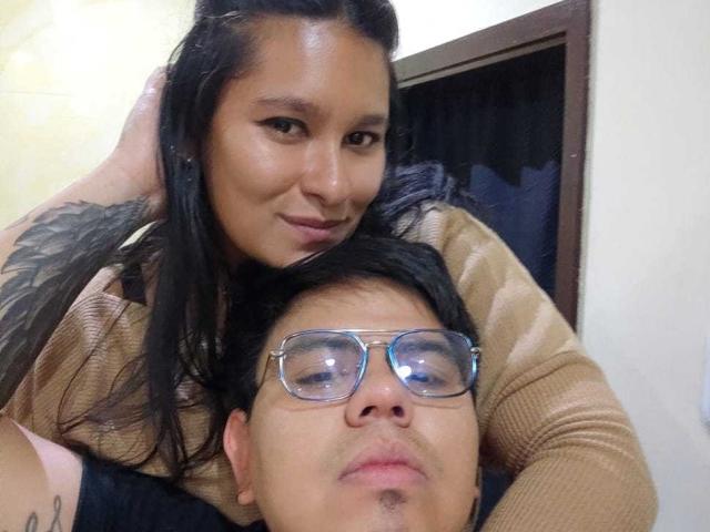 JuliaxRosee with black hair, tan sweater, and tattoo, leans over a guy with glasses, brown shirt, and black hair, smiling at the camera. His nose is slightly up.
