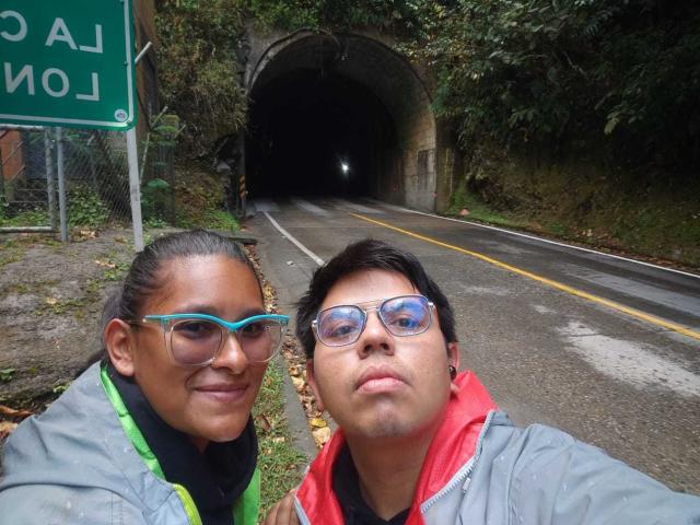 JuliaxRosee with teal glasses, brown skin, and black hair, and JuliaxRosee with glasses, short black hair, and a red jacket, standing in front of a tunnel on a road.