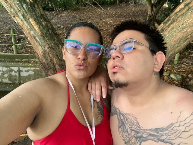 JuliaxRosee with teal glasses and red top, puffy lips, and a tattoo, stands next to a shirtless JuliaxRosee with glasses, tattoos, and a goatee, both looking at camera.