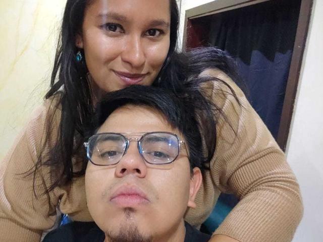 JuliaxRosee with long black hair, tan sweater, and blue earrings, leans over a guy with glasses, short black hair, and a goatee, both smiling. Her tits are slightly visible.