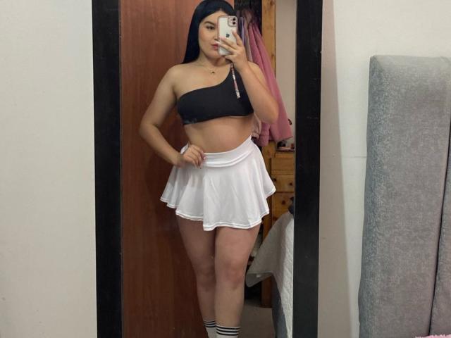 AliceMillerlu with long black hair, black one-shoulder top, white mini skirt, and knee-high socks, standing in front of a mirror, showing her tits and ass.