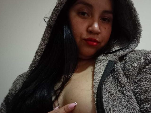 LauraMr. with dark hair, red lipstick, and a fluffy gray hooded jacket, exposing her small tits with a pink-nail-painted hand covering her nipple.