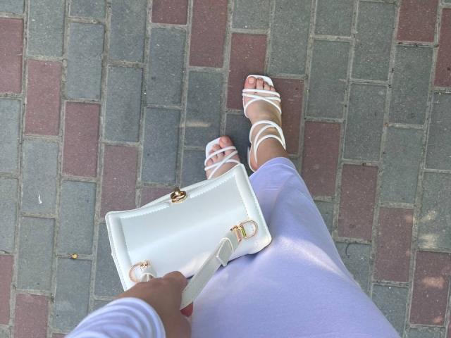 Rorria's white strappy sandals on gray and red brick pattern, holding a white clutch with gold accents, wearing light purple pants, and white sleeves, from a top-down view.