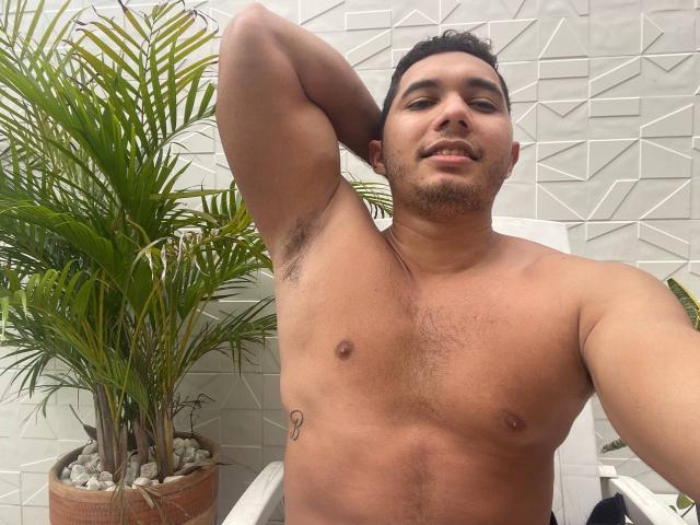 Hombre sin camisa, con un tatuaje pequeño en el pecho, brazo levantado mostrando vello axilar, cabello corto y oscuro, y una ligera sonrisa. Planta en maceta a la izquierda..