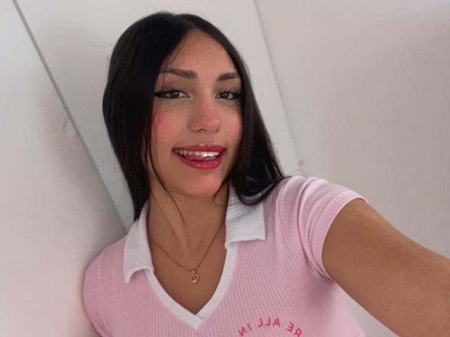 EmmaRubis with long black hair, pink ribbed shirt, white collar, red lipstick, smiling, wearing a gold necklace, holding the camera, soft white background, selfie, attractive, cute.