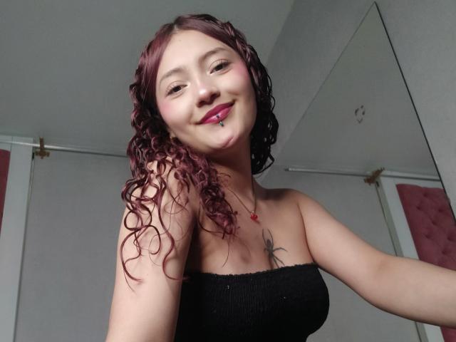 EmilyLorenz with curly red-pink hair, black strapless top, red lipstick, nose ring, and tattoo on chest, smiling at camera. Boobs visible. Mirror in background.