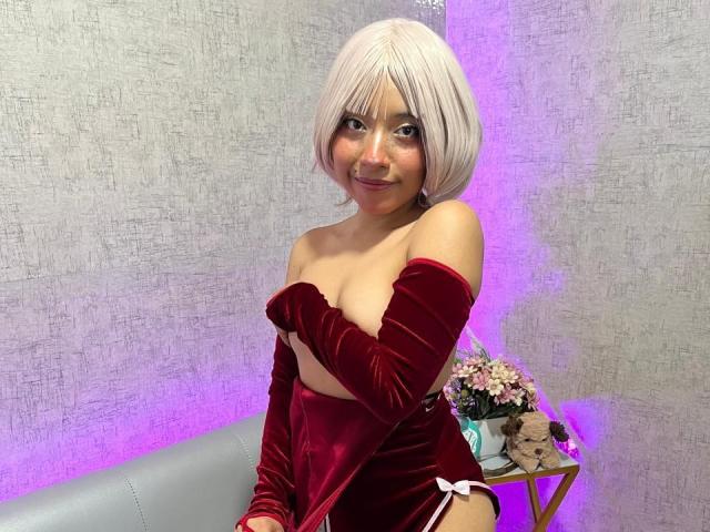 MelodyMoonz with pale pink bob, wearing red velvet off-shoulder top, exposing small tits, covering left breast, sitting, smiling, purple light, flowers in background, grey couch.
