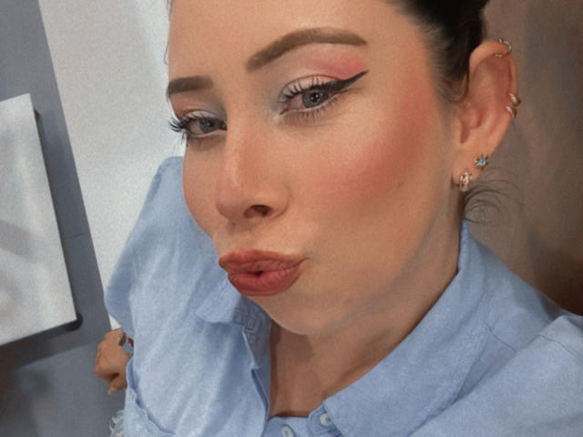 JennyLawry with puffy pink lipstick, blue eyes, and winged eyeliner, making a puckered face. She's wearing a light blue shirt and has small star earrings.