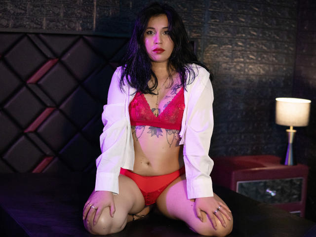 SamStormX kneels on a bed wearing red lace bra and panties, with a white shirt open, showing her tits and pussy. Dark wavy hair, tattoos on her chest.