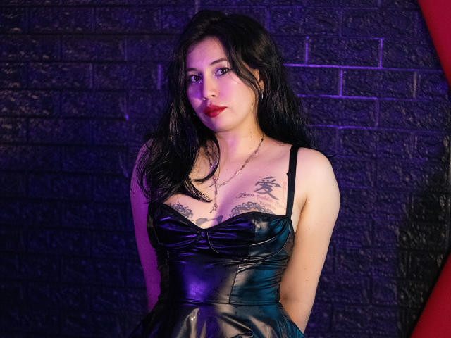 SamStormX with black leather dress, showing tits with tattoos, standing against brick wall, purple and blue lighting, long black hair, red lipstick, hands behind back, looking at camera.