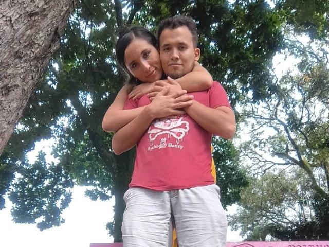 HinataAndNoha, a woman with long black hair, hugs a man with short brown hair, wearing a pink shirt and white pants, standing under a tree. She's grabbing his tits.