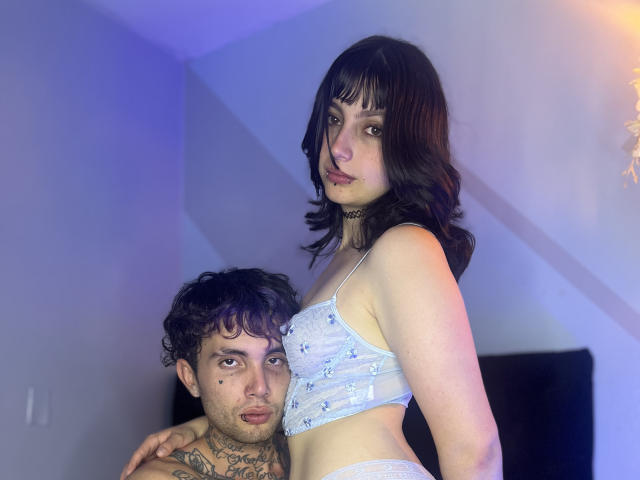 TwoNightmares with dark wavy hair, white lace bra, and choker, stands beside a curly-haired dude with tattoos, wearing only white lace panties, in a dimly lit room.