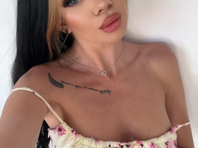 IvyRaven with black and blonde hair, wearing a floral dress, showing her tits, a crescent moon tattoo, and a small cross necklace, with pink lips and a nose ring.
