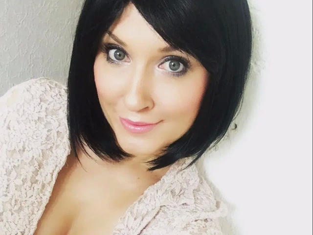 MissDoertie, con un corte de pelo bob negro, ojos grises, lápiz labial rosa y una blusa de encaje, sonriendo a la cámara, mostrando ligeramente sus senos. Fondo blanco y suave, pared con textura..