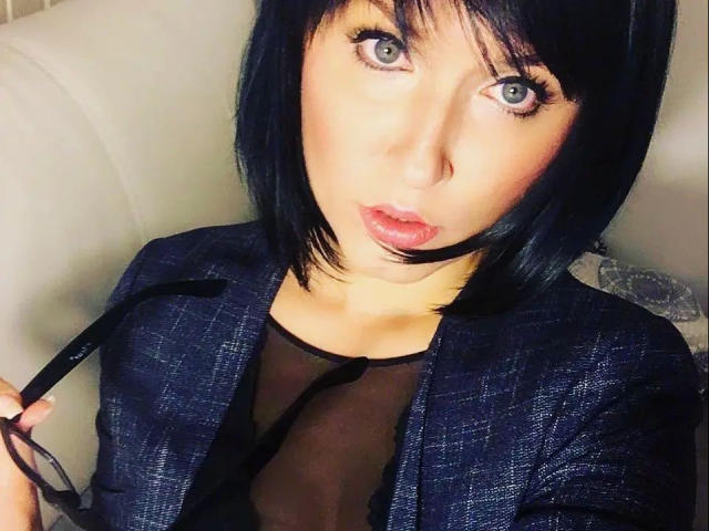 MissDoertie, con un corte de pelo bob negro, sosteniendo gafas, vistiendo un blazer texturizado azul oscuro, una blusa negra transparente y lápiz labial rosa, mirando hacia arriba con ojos azules grandes y labios fruncidos..