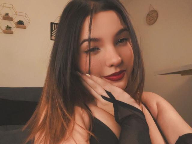 MiaCastillos with long brown hair, red lipstick, black lingerie, and fingerless black gloves, smiling at the camera, hands on cheek. Her tits are visible in the black top.