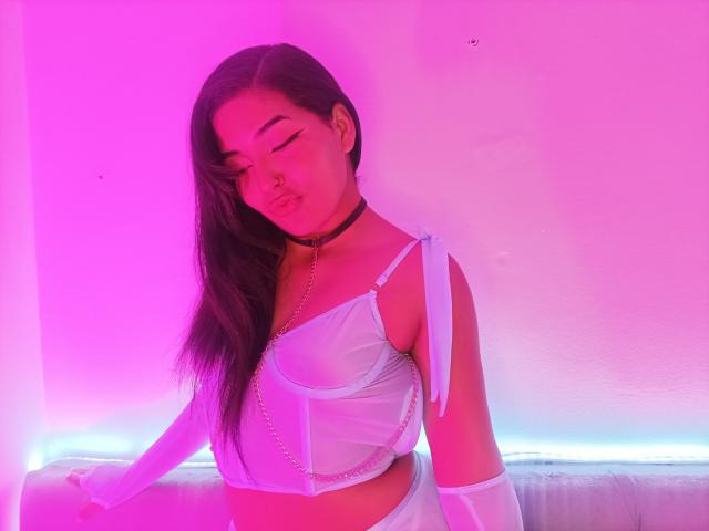 RebecaGomez with long black hair, white lingerie, and chain choker, pink and purple neon light, eyes closed, pouty lips, standing, white wall, white couch, arm resting.