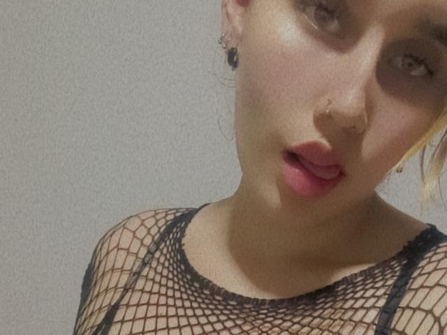 Keidy69 with pouty pink lips, blonde hair, and a black fishnet top, showing her tits through the net. She has a nose piercing and small ear rings.